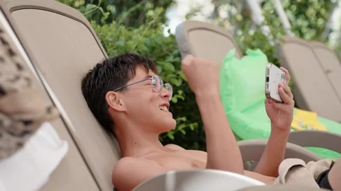 Teen boy using smartphone on sun lounger at summer resort pool Stock Footage 336116201