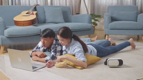 Teen Couple Using A Laptop While Lying On Carpet On The Floor At Home. Eating Stock Footage 247975478