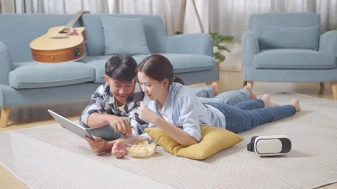 Teen Couple Using Tablet While Lying On Carpet On The Floor At Home. Eating Stock Footage 247975778