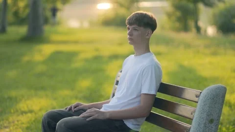 Teen guy in white tee chilling solo on stone bench in green city park Stock Footage 315036730