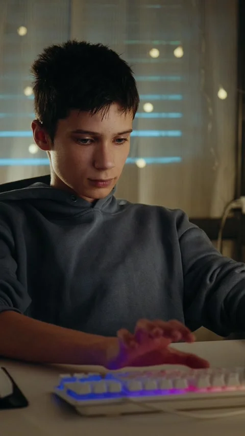 Teenage boy intently typing on a computer with an illuminated keyboard at night. Stock Footage 336316936