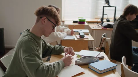 Teenage Boy Taking Notes at Single-Seat Desk Among Other Students in Class Stockbeeldmateriaal 335690160