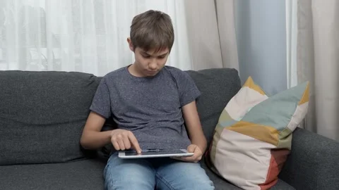 Teenage boy using digital tablet computer sitting on couch at home. Kid using Stock Footage 165988588