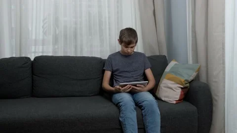 Teenage boy using digital tablet computer sitting on couch at home. Kid using Vídeo Stock 166038130