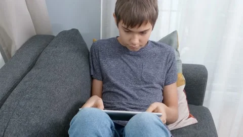 Teenage boy using digital tablet computer sitting on couch at home. Kid using Stock Footage 166163053