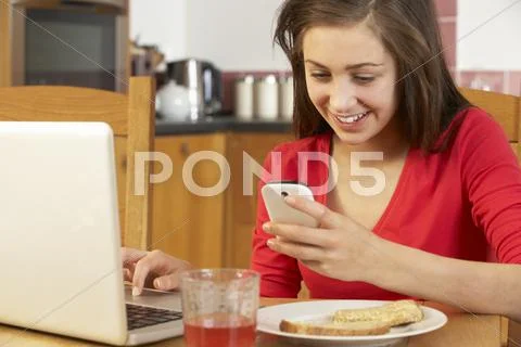 Teenage Girl Using Laptop And Mobile Phone Whilst Eating Breakfast Stock-Fotos