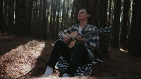 Teenager leans back on tree while playing acoustic guitar in the forest. Stock Footage 142299333