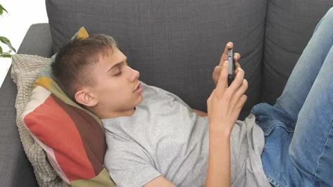 Teenager lying on sofa using smartphone. Boy is sick, coughs. Kid watching video Stock Footage 174167320