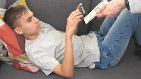 Teenager lying on sofa using smartphone, gaming. Mom gives him 5 dollar banknote Stock Footage 177356485