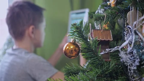 A teenager is playing with a tablet while sitting behind a Christmas tree. New Stock Footage 166852820