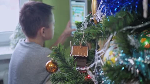 A teenager is playing with a tablet while sitting behind a Christmas tree. New Stock Footage 167325634
