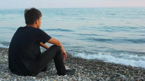 Teenager sits on pebble beach and looks at sea, back view Stock Footage