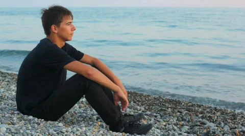 Teenager sits on pebble beach and looks at sea, profile Stock Footage