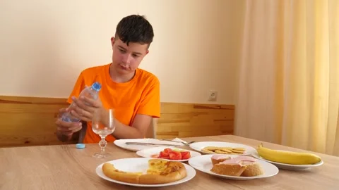 A teenager sits at a table, eats and drinks water. Stock Footage 319985174