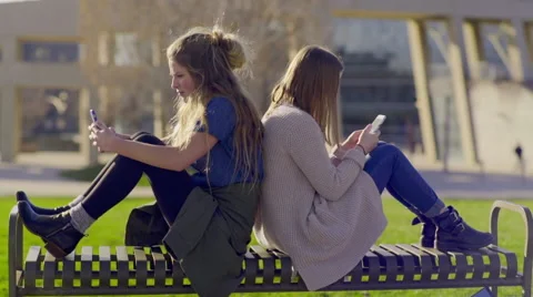Teens Sit Back To Back On A Park Bench And Check Their Smartphones Stock Footage 61842013