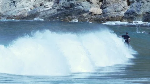 Telephoto Surfers riding a wave in a day of sun near a rocky beach Stock Footage 89581663