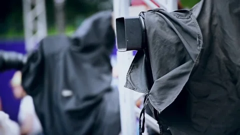 Television news camera on a tripod protected by a black rain cover Видео 78098691