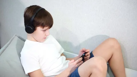 Ten year old boy lying on the couch playing on smartphone with wireless headphon Stock Footage 297553363