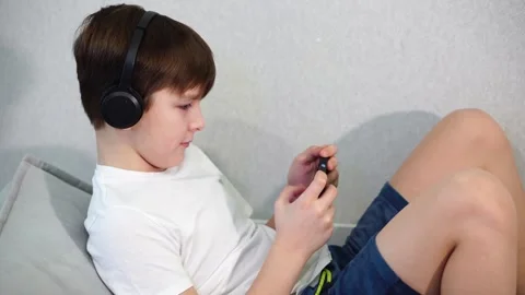 Ten year old boy lying on the couch playing on smartphone with wireless headphon Stock Footage 297553460