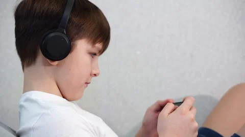 Ten year old boy lying on the couch playing on smartphone with wireless headphon Stock Footage 297553462