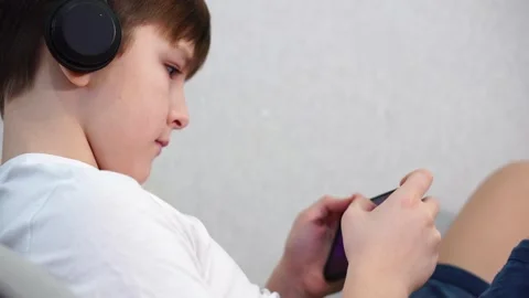 Ten year old boy lying on the couch playing on smartphone with wireless headphon 库存影片 297553519