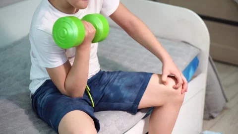 Ten year old boy pumping biceps with green dumbbell sitting on bed. Stock Footage 307023131