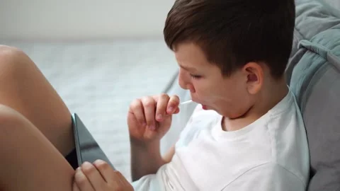 Ten year old boy sitting on bed watching video on tablet and sucking lollipop Stock Footage 307022870