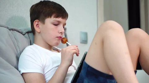 Ten year old boy sitting on bed watching video on tablet and sucking lollipop 库存影片 307022872