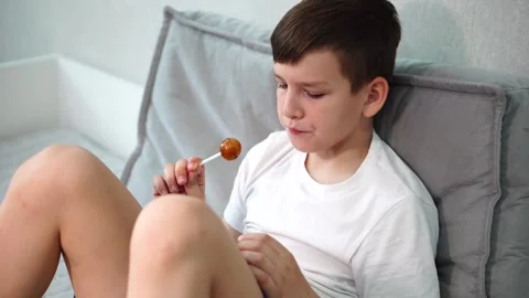 Ten year old boy sitting on bed watching video on tablet and sucking lollipop Stock Footage 307022899