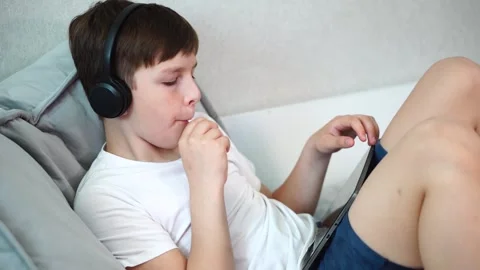 Ten year old boy sitting on bed with headphones watching video on tablet and suc Stock Footage 307022960