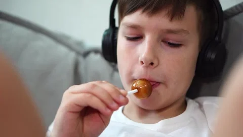 Ten year old boy sitting on bed with headphones watching video on tablet and suc Stock Footage 307023025