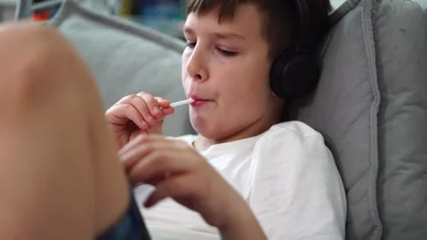Ten year old boy sitting on bed with headphones watching video on tablet and suc Stock Footage 307023026