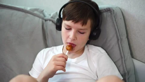 Ten year old boy sitting on bed with headphones watching video on tablet and suc 库存影片 307023049