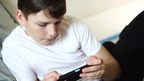 A ten-year-old boy in a white T-shirt enthusiastically plays on his phone while  Stock Footage 306445382