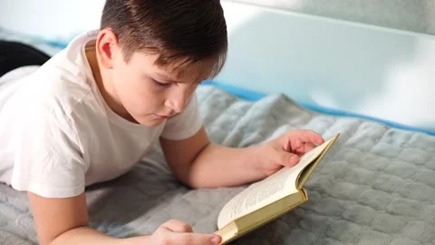 Ten year old boy in white t-shirt lying on bed reading hardcover book Stock Footage 306445501