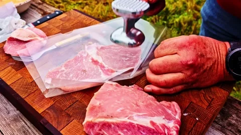 Tenderizing raw pork cutlets with meat mallet on a wooden butcher block outdoors Stock Footage 325570521