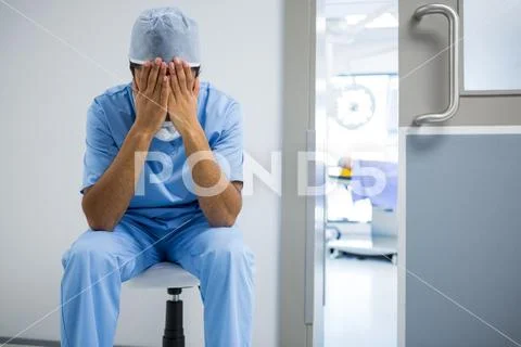 Tensed surgeon sitting with hands on face in corridor Stock Photos