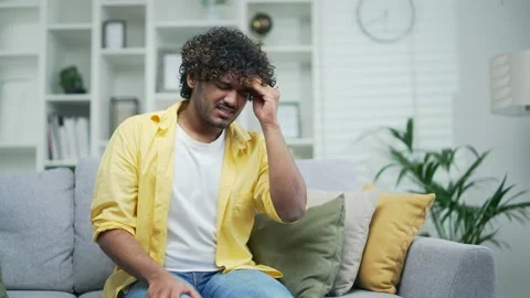 Tension and headache in a young handsome man sitting at home on the couch. Curly Stock Footage 235488045