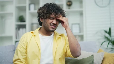 Tension and headache in a young handsome man sitting at home on the couch. Curly Stock Footage 235488093
