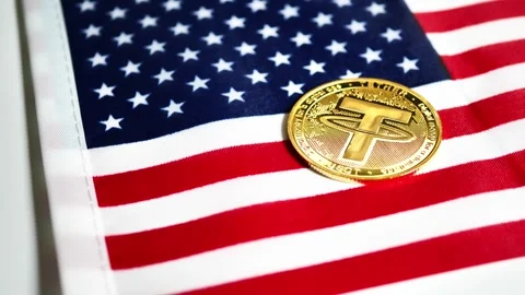 A Tether (USDT) cryptocurrency coin is positioned on an American flag Stock Footage 316233638