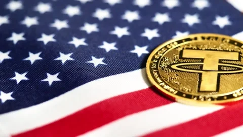 A Tether (USDT) cryptocurrency coin is positioned on an American flag Stock Footage 316233692