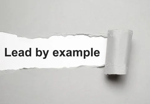 The text lead by example appearing behind torn grey paper Stock Photos