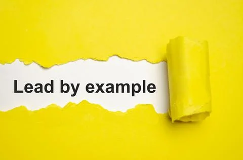 The text lead by example appearing behind torn grey paper Stock Photos