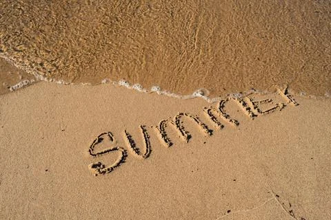 Text SUMMER handwritten in sand surface. Blue ocean wave washing away message at Stock-Fotos