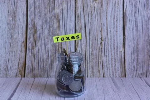 Text on yellow note on a jar full of coins on wooden desk. Taxes Stock Photos
