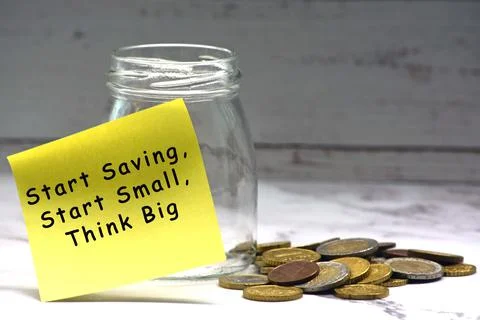 Text on yellow sticky note with blurred glass jar and coins on table surface Stock Photos