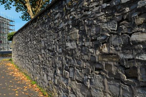 A textured stone wall alongside a tree-lined street in autumn Stock Photos