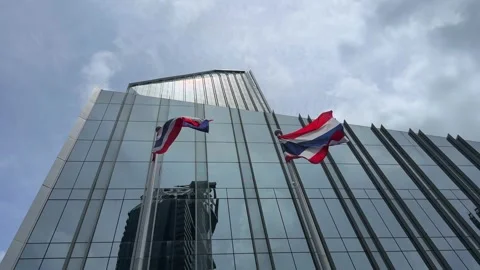 Thai flags waving in front of modern glass skyscraper reflecting city skyline 스톡 동영상 314354497