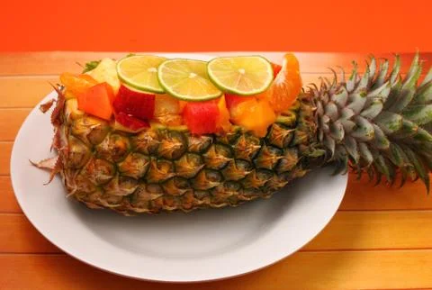 Thai style fruit salad inside a pineapple with an orange background on a wood ta Stock Photos