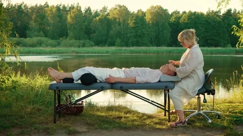 Therapist gently massaging face of client in open air setup. Calm lake and Stock Footage 301994503